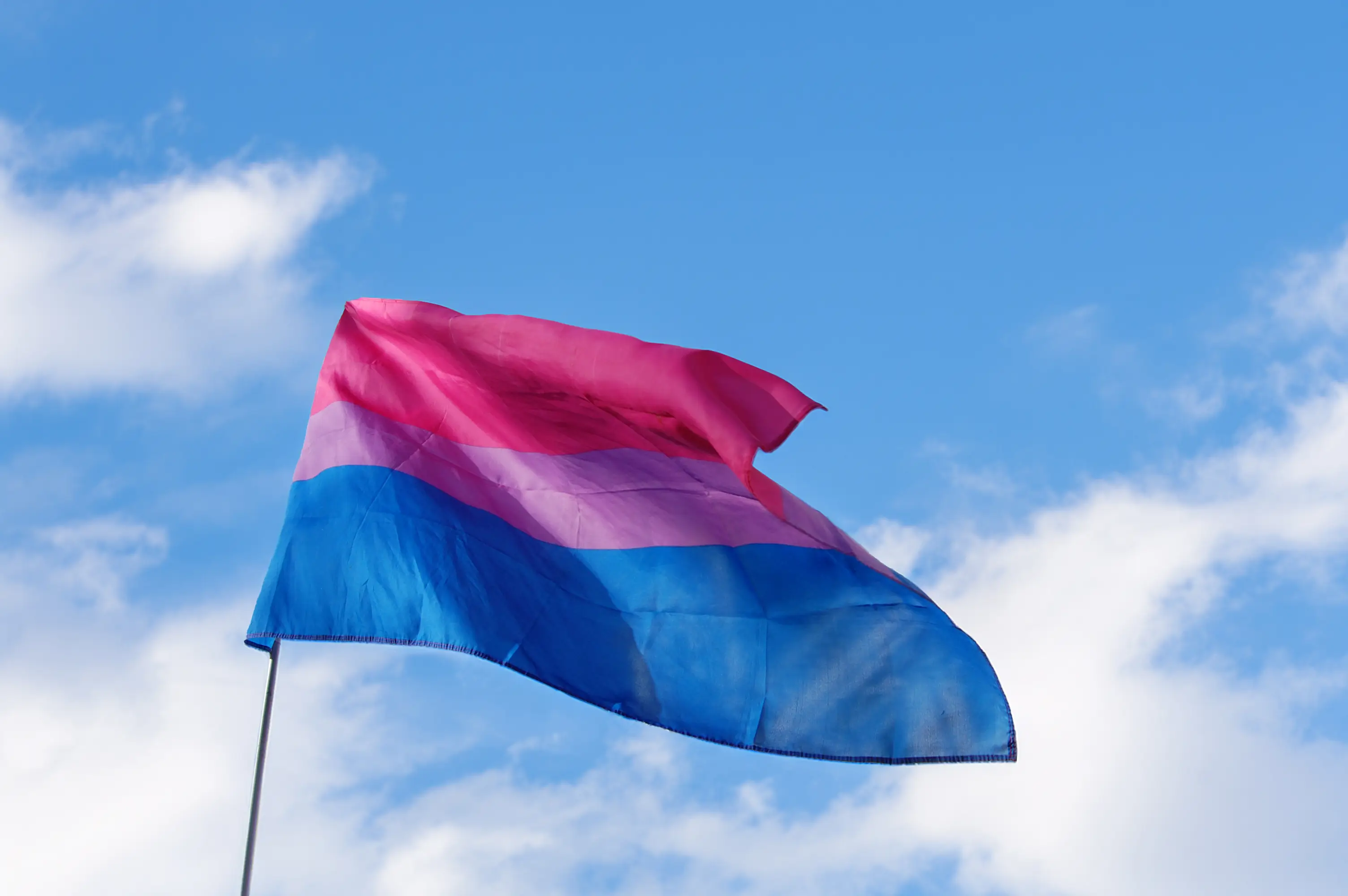 Wehende Bi+-Flagge in Rosa, Lila und Blau vor blauem Himmel mit leichten Wolken